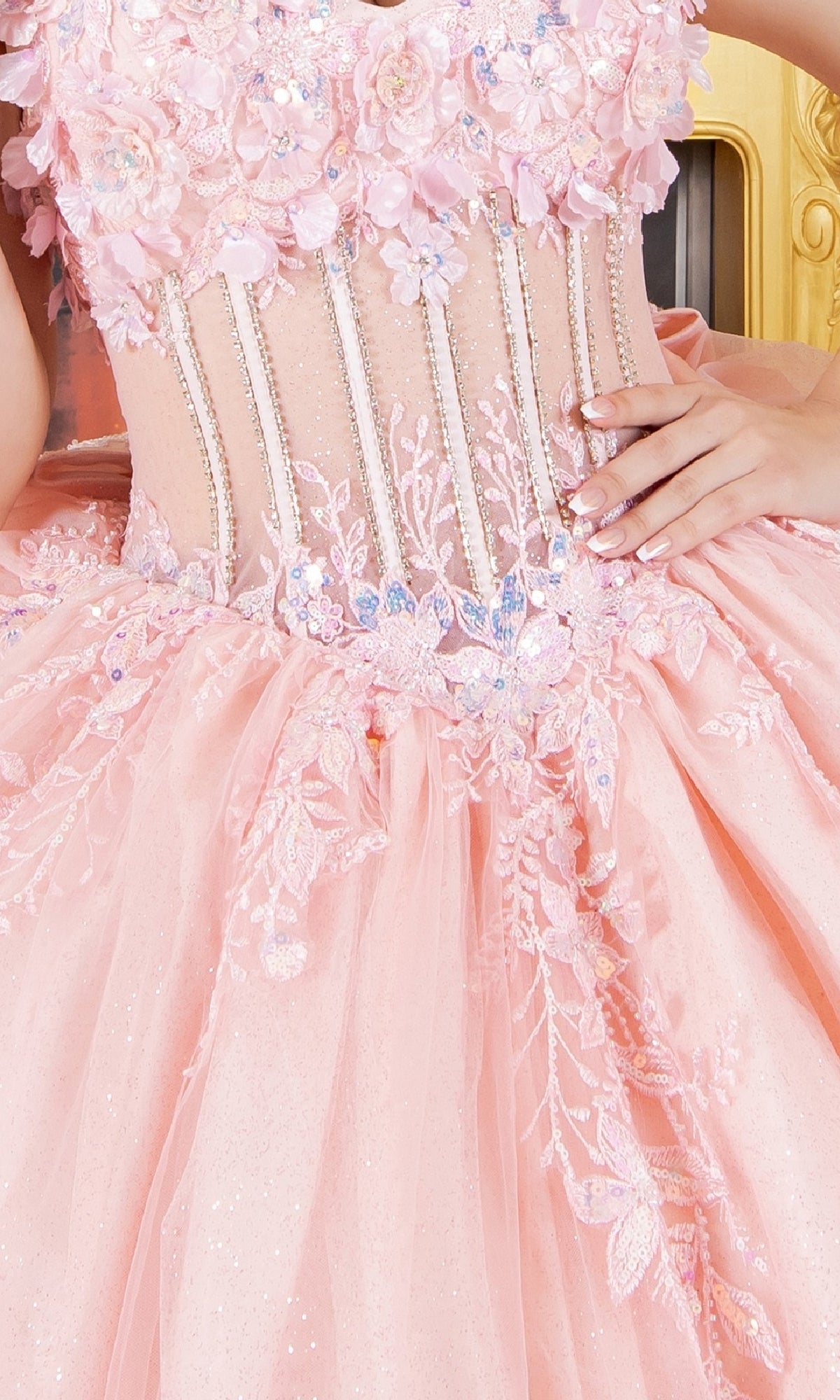 A person wearing the Cinderella Couture 8160J quinceanera dress, pink and off-the-shoulder with floral-embroidered lace, beaded details, and a sheer corset bodice, poses with one hand on their waist.